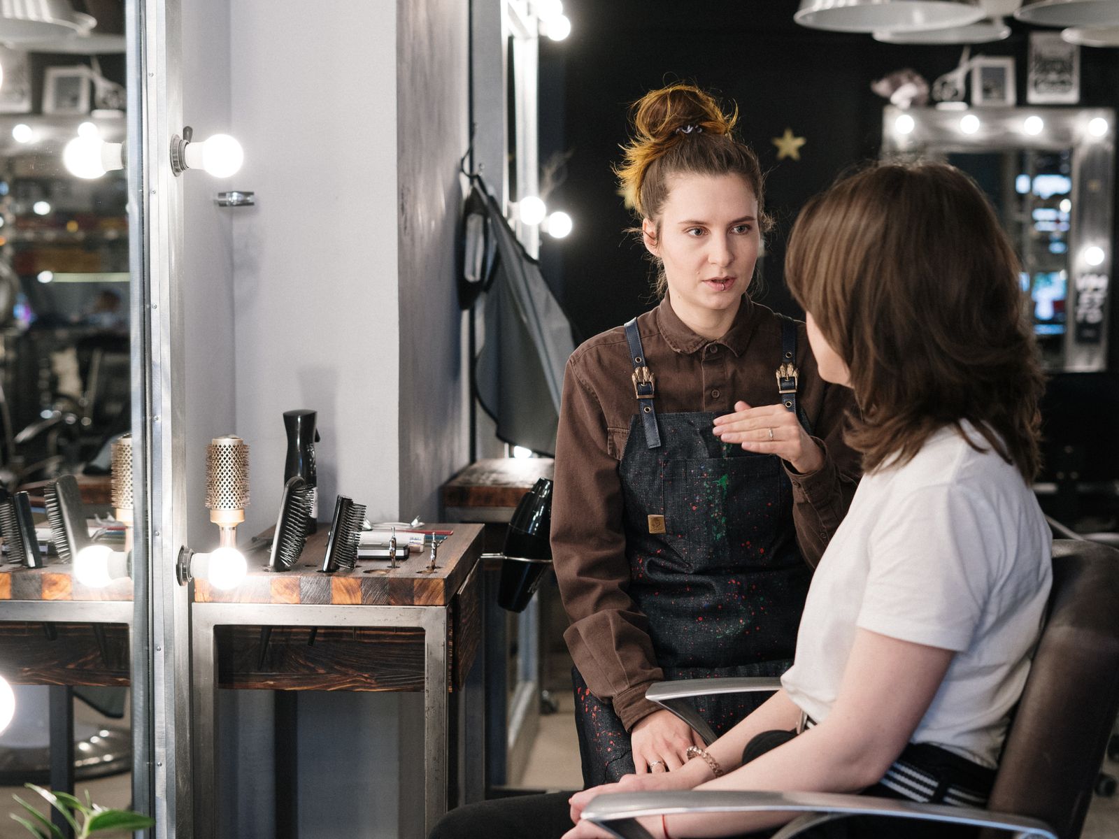 A stylist leaning in to talk with a client in her chair, mid-consultation in a small salon.